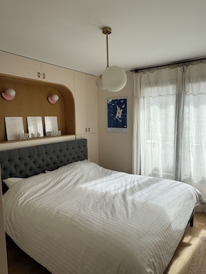 A bedroom with a tufted headboard, a bed with a white duvet, a ceiling fan, and a wall-mounted shelf with framed pictures and a map. - Modern, family-friendly Parisian apartment (Paris)