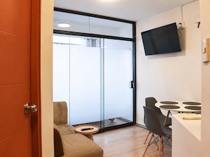 Ein modernes Zimmer mit einer Schiebetür zum Balkon, einem kleinen runden Tisch mit Fernbedienung, einem Esstisch mit Stühlen und einem an der Wand befestigten Fernseher.