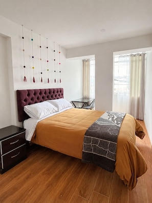 Ein Schlafzimmer mit einer gebogenen Kopfstütze, einem Bett mit braun-grauer Decke, einem Nachttisch und einem Wandbild.