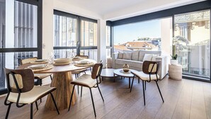 Un comedor moderno con una mesa redonda de madera, sillas y un sofá. Amplias ventanas ofrecen vistas a edificios y un cielo despejado.