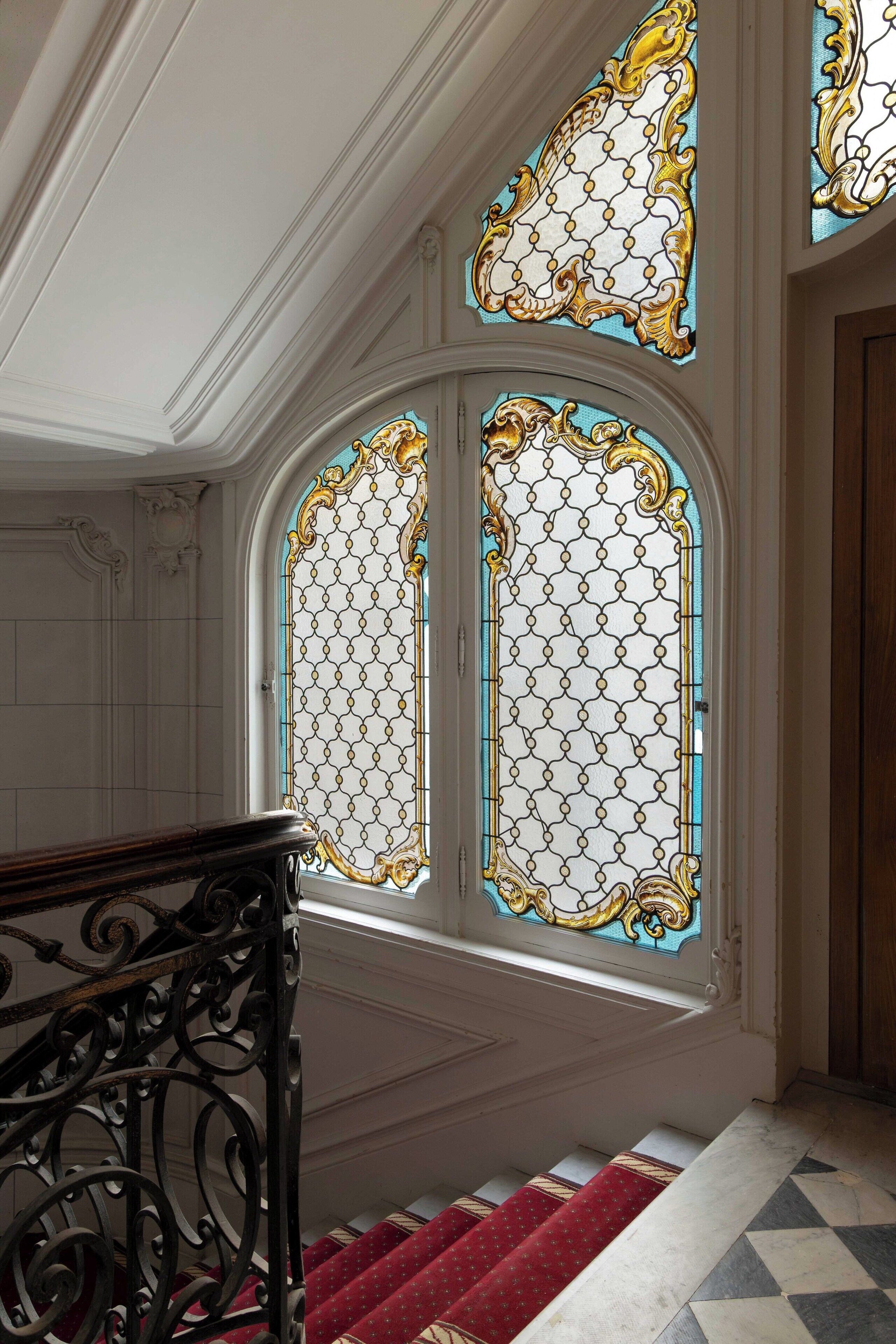 A grand staircase with ornate balustrades and a red carpet, leading to a room with large stained glass windows.