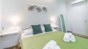 Hotellihuone, jossa on sänky, vihreät vuodevaatteet, kaksi tyynyä, seinälle ripustettu taideteos, yöpöytä lampun kanssa ja ovi, jossa on pyyheteline.
