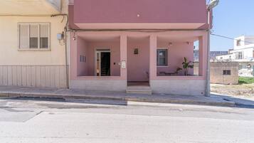 Un edificio de dos pisos con fachada rosa, una entrada visible y un pequeño espacio al aire libre con mesas.