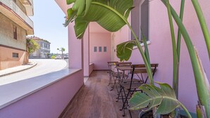 Uma varanda estreita com uma mesa e cadeiras de madeira, um vaso de planta e vista para a rua e prédios.