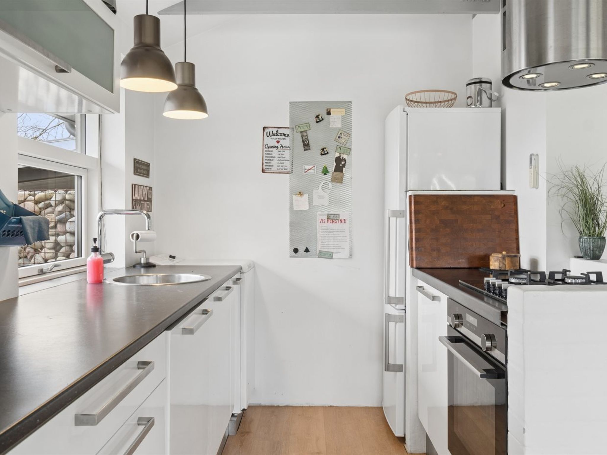 A modern kitchen with white cabinetry, stainless steel appliances, pendant lighting, and a bulletin board with various notices.