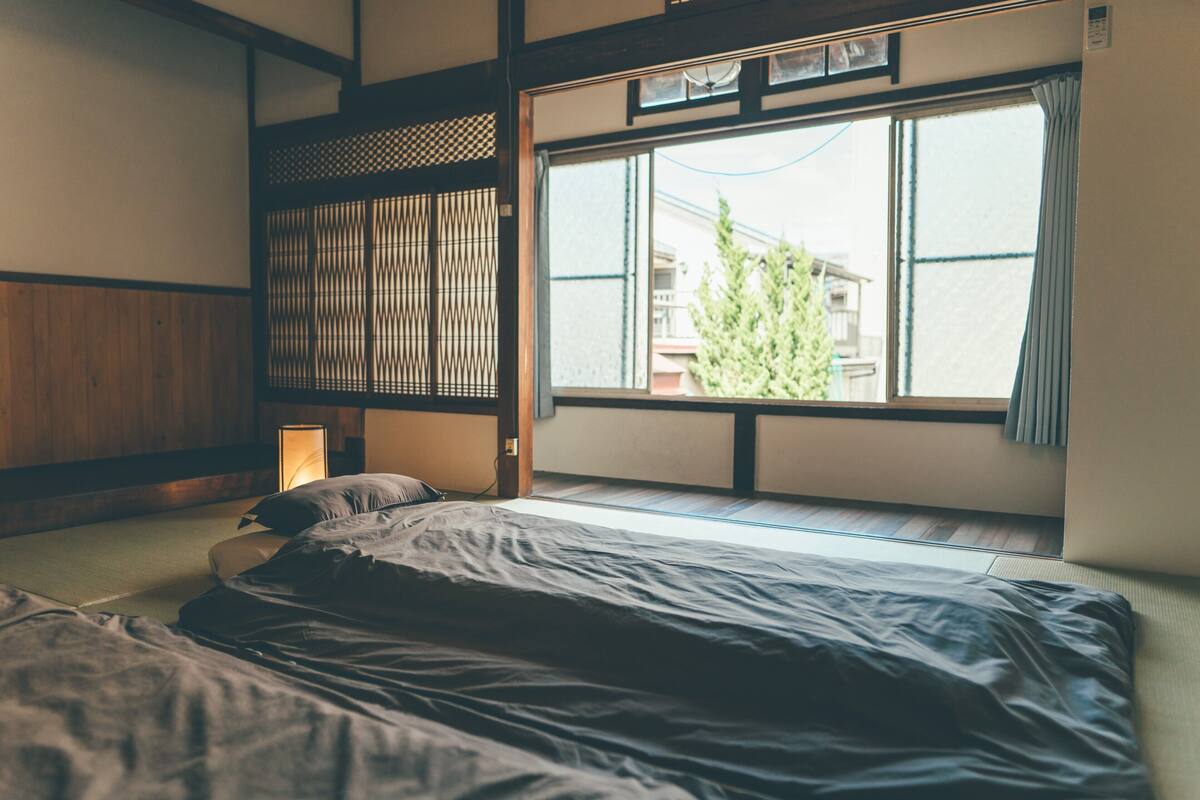 A traditional Japanese-style room with tatami flooring, a low bed, a sliding shoji screen, and a large window with a view of a building and trees.