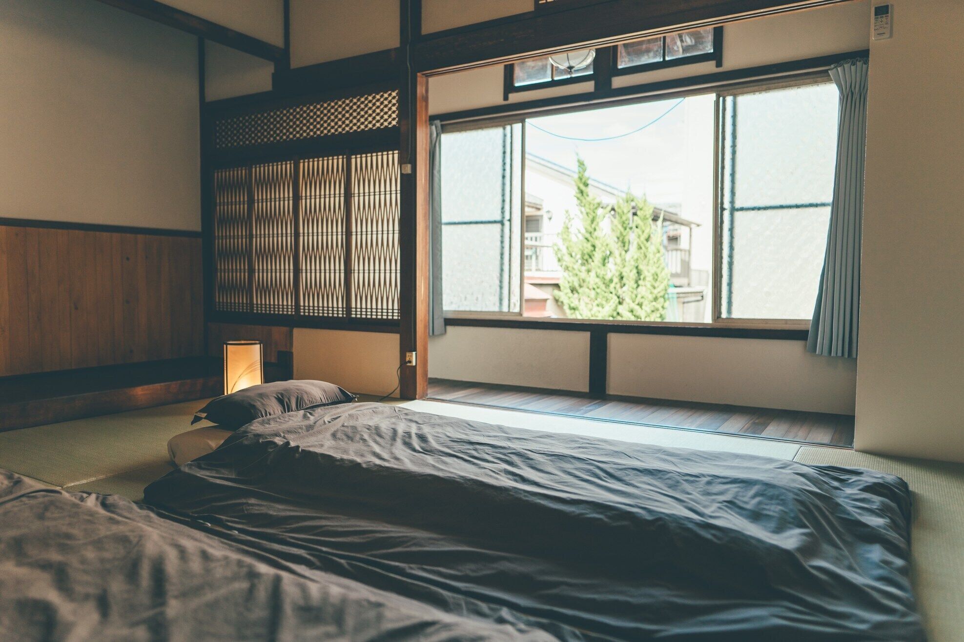 Una habitación tradicional japonesa con suelo de tatami, futón bajo, celosía de papel y un ventanal con vistas a edificios y árboles.