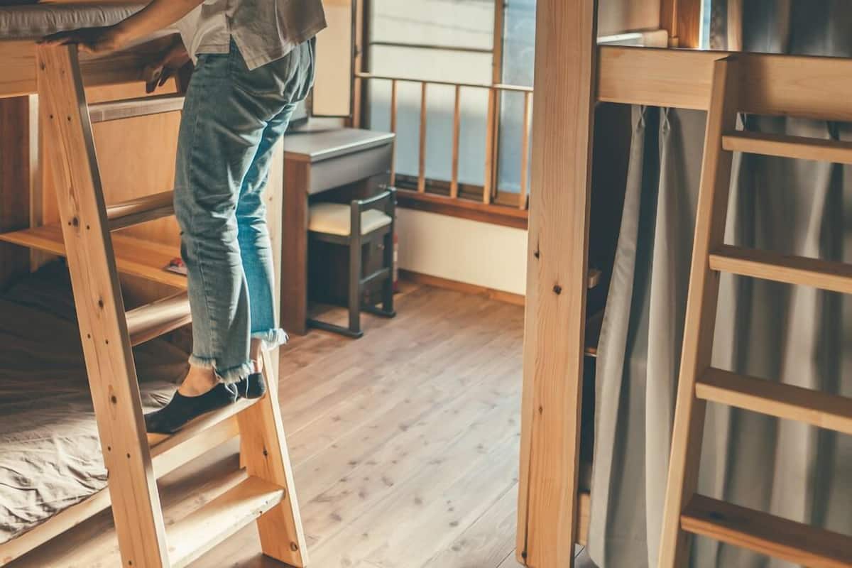 A bunk bed with a wooden ladder, a desk, and a chair in a room with wooden flooring and a window.