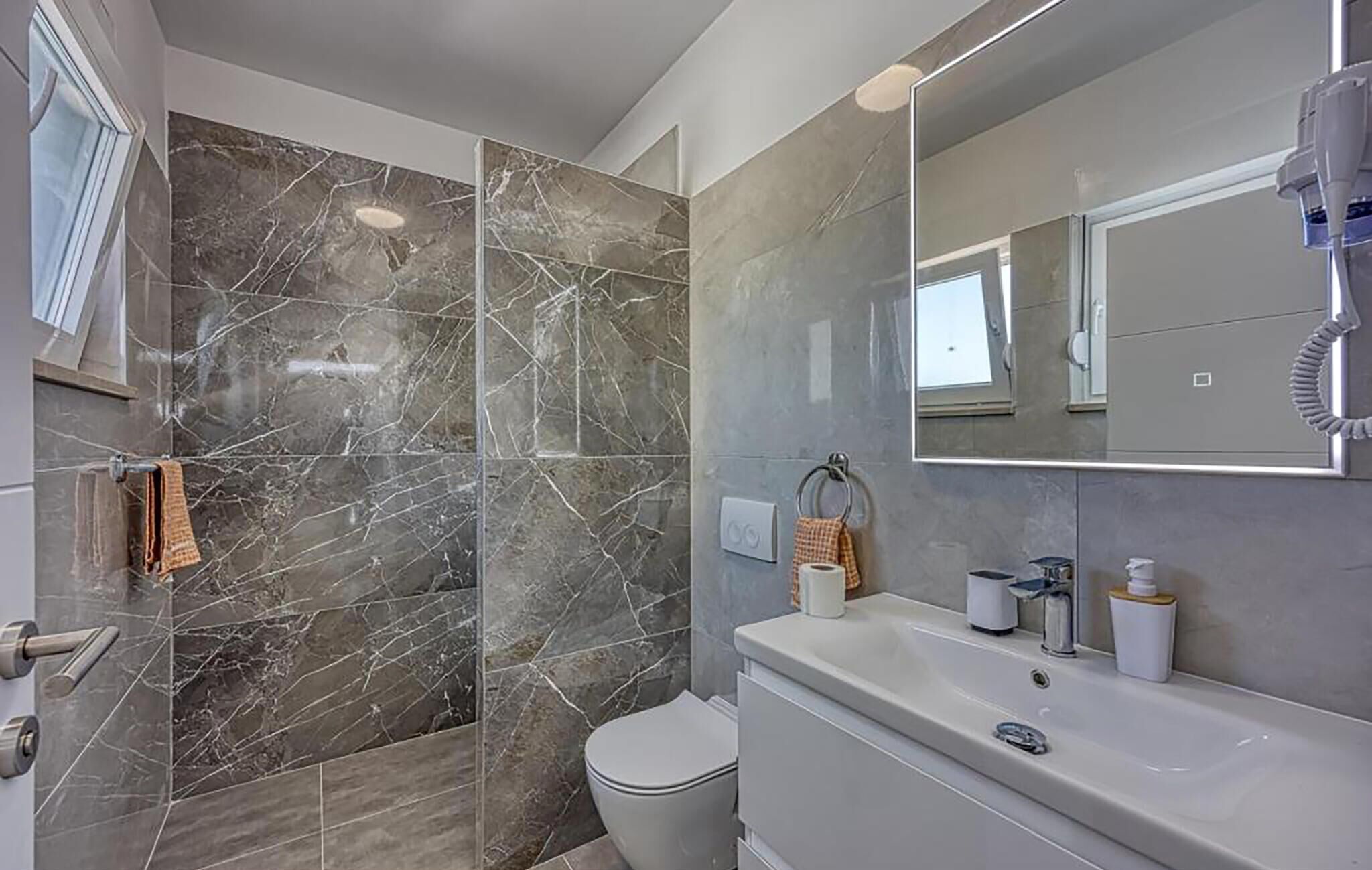 Une salle de bain moderne avec un mur de douche en marbre, des toilettes, un lavabo avec des articles de toilette et un grand miroir.