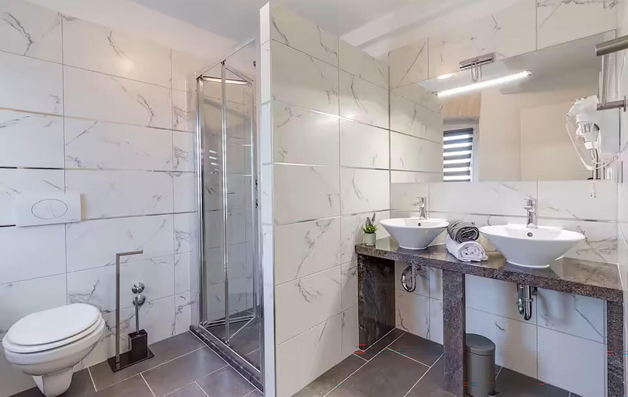 A modern bathroom with a glass shower enclosure, a toilet, and two sinks with marble countertops.