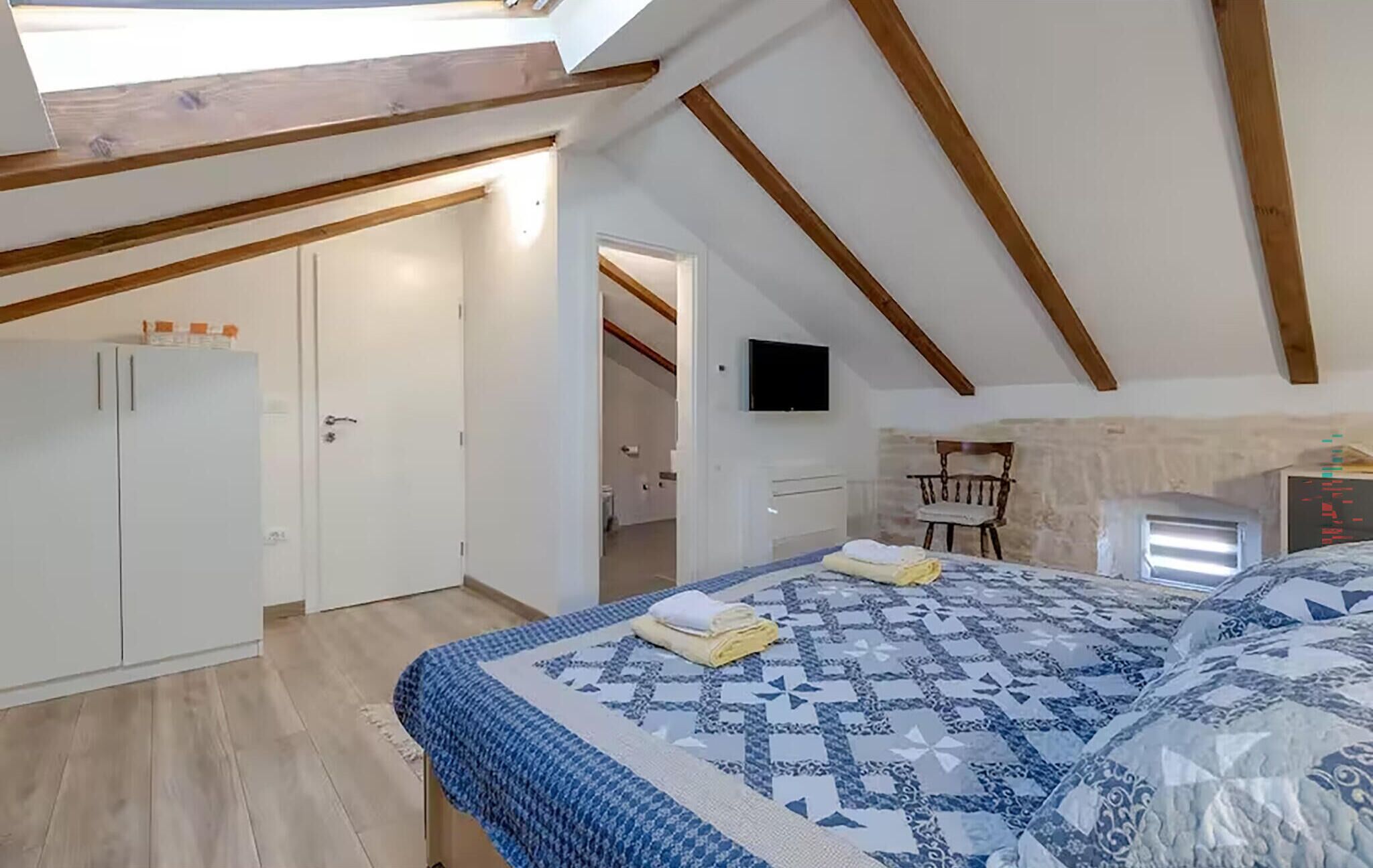 Un dormitorio con techo inclinado, una cama con colcha estampada, un televisor montado en la pared y una silla de madera.