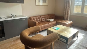 A modern living room with a brown leather sofa, a wooden coffee table, a kitchen area with grey cabinets, and a framed picture on the wall. - 3-bedroom apartment in the heart of the Alpes Mancelles, 20 minutes from the 24h of Le Mans (Beaumont-sur-Sarthe)