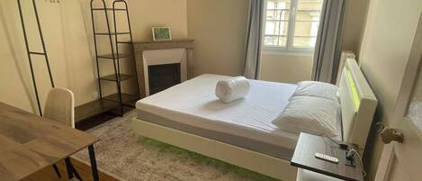 A bedroom with a bed, a desk, a chair, a radiator, a window with curtains, and a small fireplace.