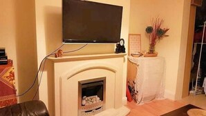 Een woonkamer met een open haard, een flatscreen televisie erboven, een kleine tafel met een vaas, en een bruine leren bank.