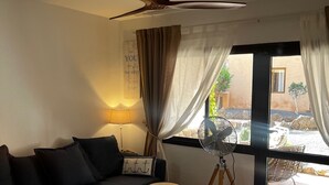 A cozy living room with a dark sofa, a small round table, a fan, and a view of the outside. - Susana - cozy  1-bedroom apartment in charming Corralejo perfect for relaxing (Corralejo)