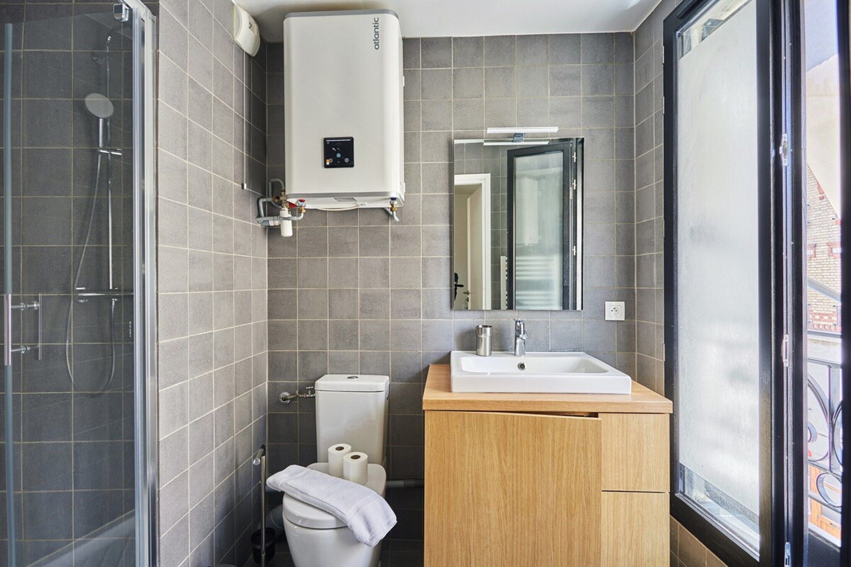A modern bathroom with a glass shower enclosure, a toilet, a sink with a mirror above it, and a window providing a view of the outside.