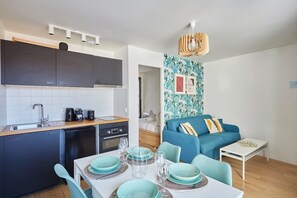 A compact living space with a kitchenette, dining area, and a living room with a blue sofa and patterned wallpaper. - Charming Apartement Boulogne Billancourt Center (Boulogne-Billancourt)