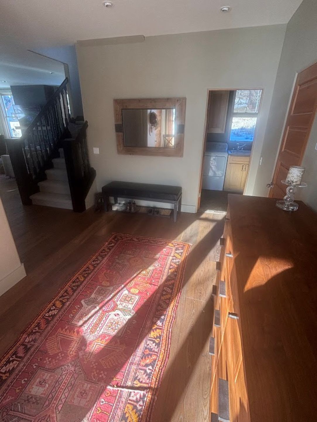 A spacious living area with a wooden floor, a red patterned rug, a bench, and a kitchen visible through an open door.