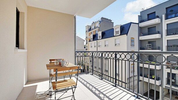 Un balcón con mesa y sillas de madera, barandilla de metal y vistas a edificios cercanos.