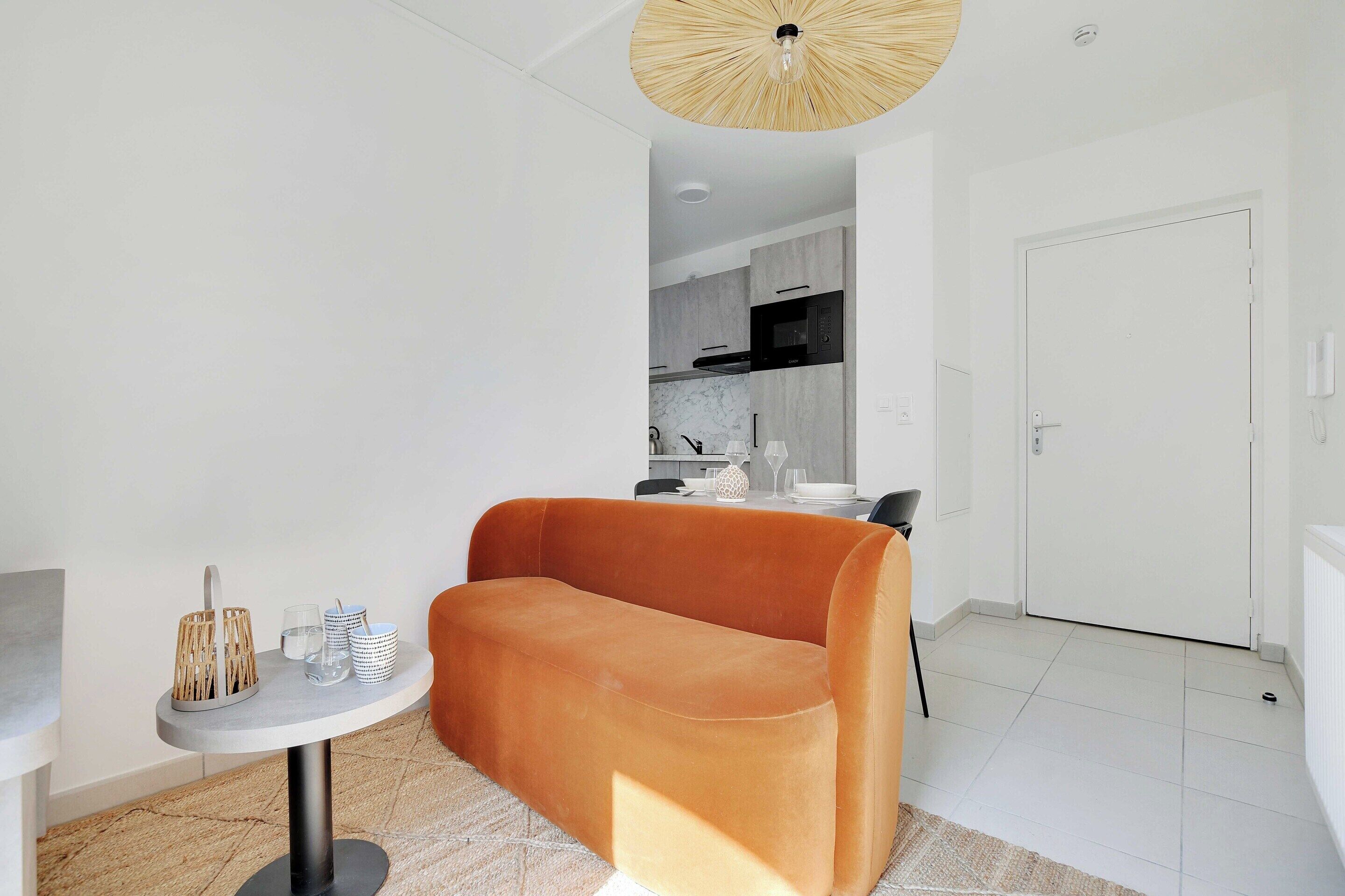 Una sala de estar moderna con un sofá, una mesita redonda con una lámpara, y una cocina al fondo.