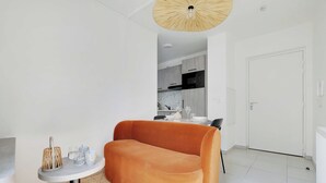 Una sala de estar moderna con un sofá, una mesita redonda con una lámpara, y una cocina al fondo.