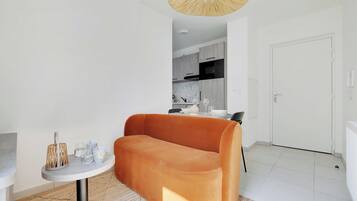 Una sala de estar moderna con un sofá, una mesita redonda con una lámpara, y una cocina al fondo.