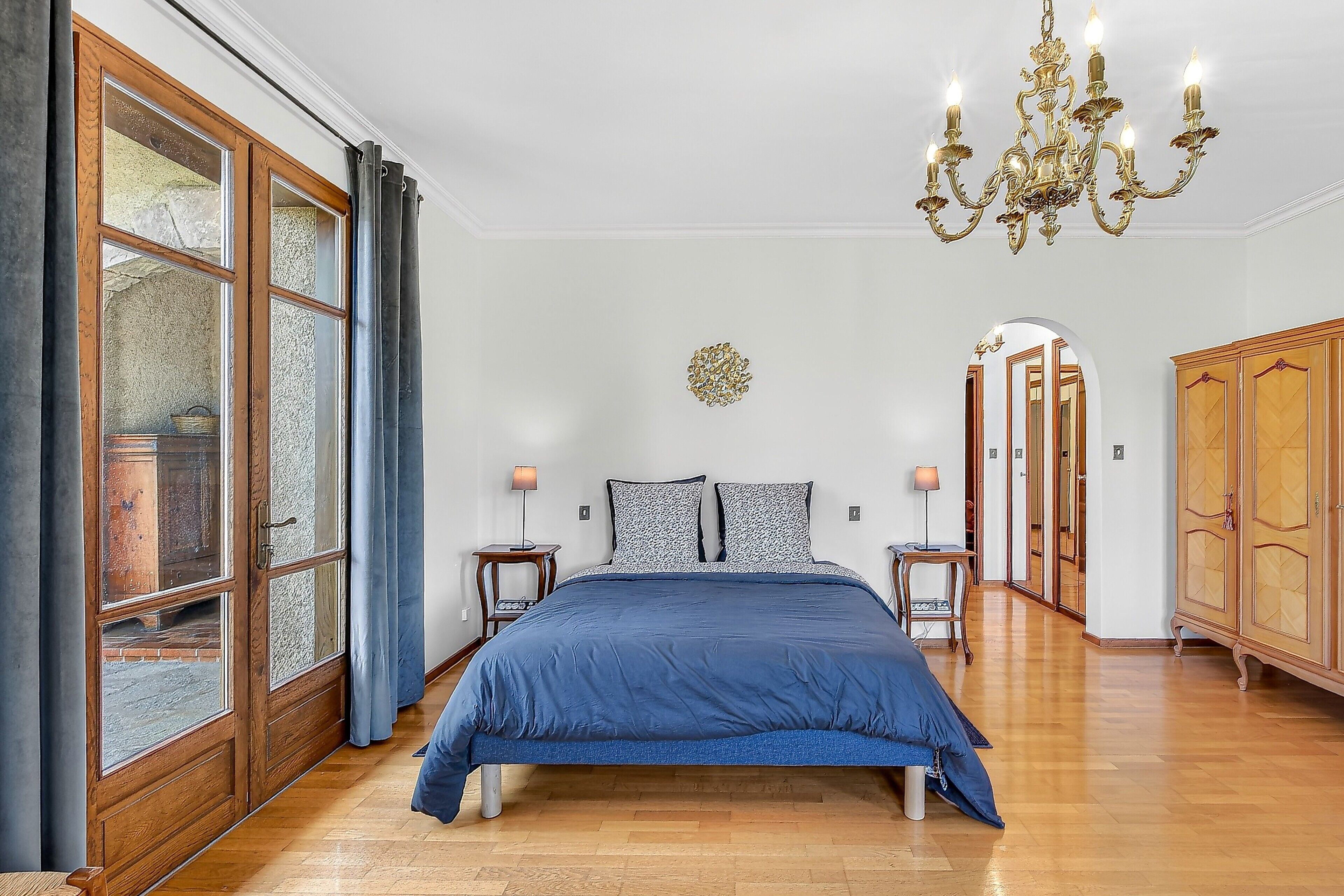 Une chambre à coucher avec un grand lit, un lustre, une armoire en bois et une vue sur l’extérieur par une fenêtre ornée de rideaux.