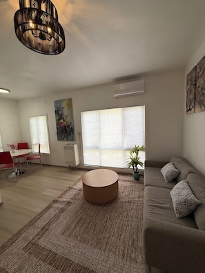 Ein modernes Wohnzimmer mit einem runden Couchtisch, einer Couch, einem Essbereich mit roten Stühlen und einem großen Fenster mit Jalousien.