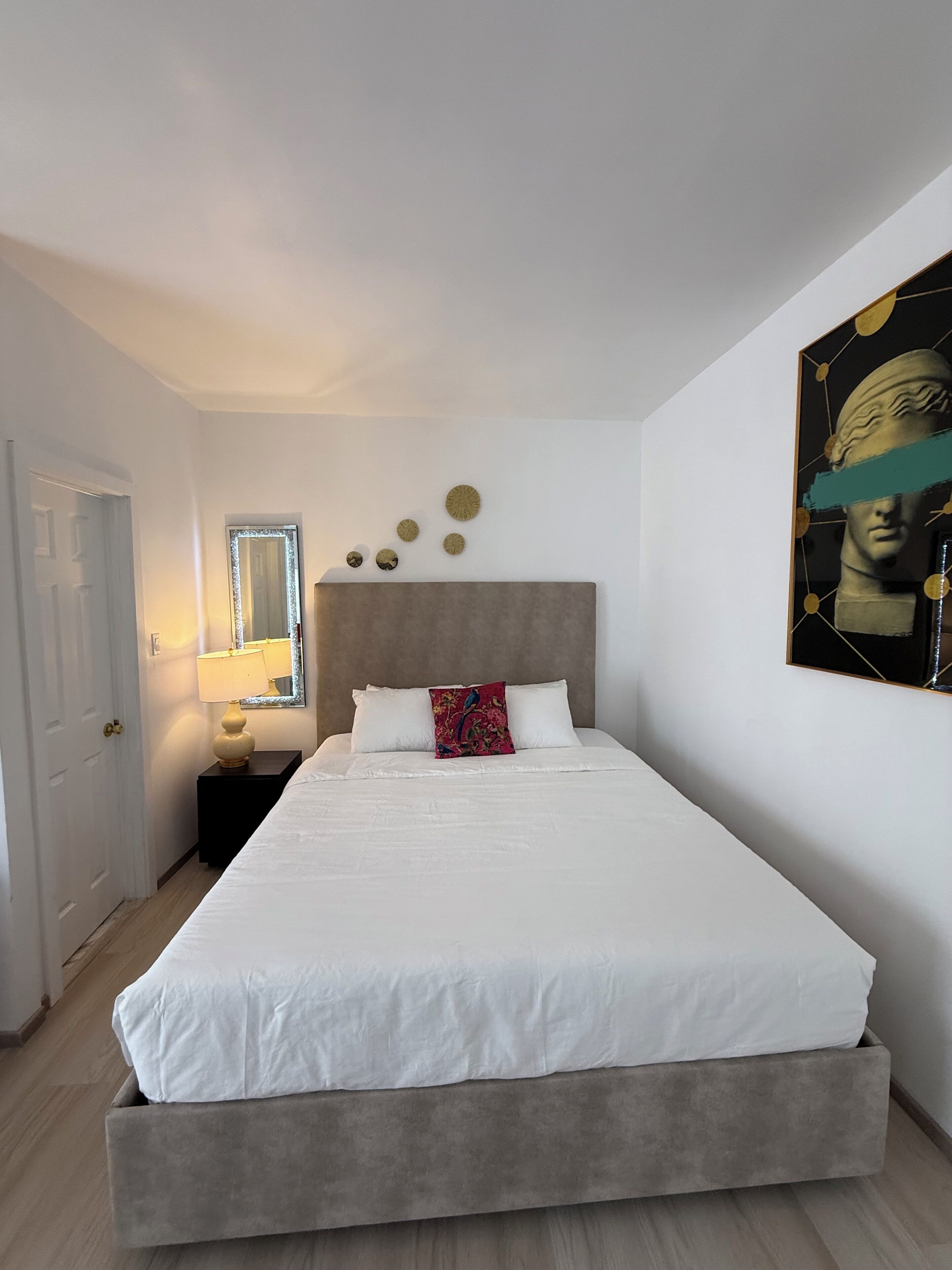 Quarto com cama, mesa de cabeceira, luminária, espelho e quadros nas paredes.