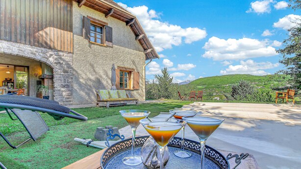 A terrace with a table set for drinks, a hammock, and a view of a house with stone walls and wooden shutters. - Le Domaines Des Thermes – T2 with Shared Pool, Peaceful Setting, Mountain View (Gap)