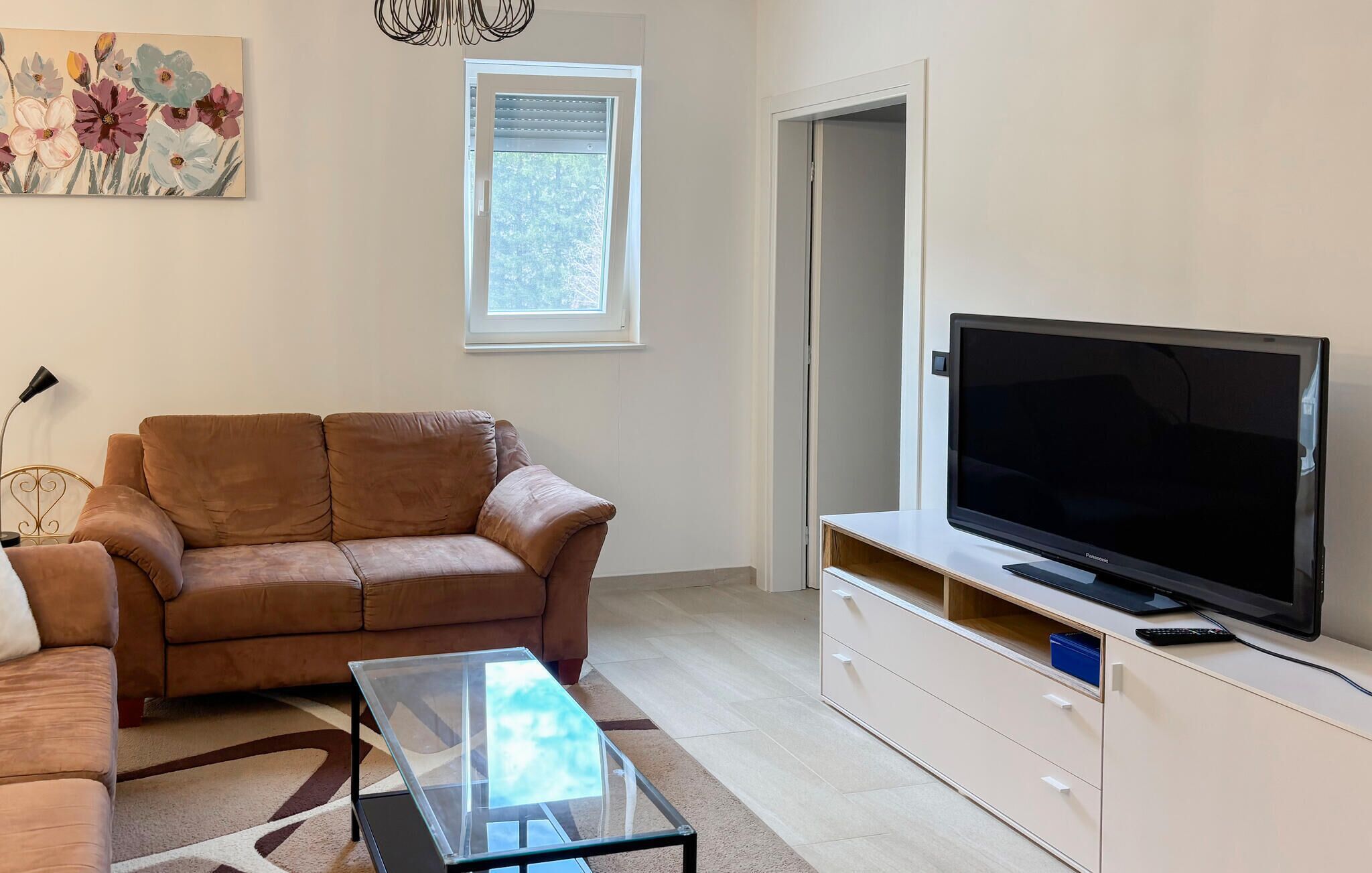 Ein Wohnzimmer mit braunem Sofa, einem Glastisch, einem Flachbildfernseher auf einer weißen Kommode, einem Bild an der Wand und einem Fenster mit Jalousien.