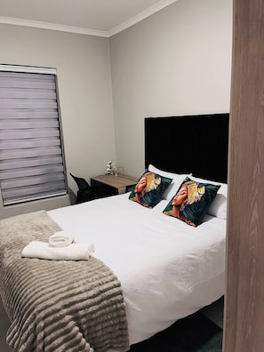 A hotel room with a bed, a desk, a chair, a window with blinds, and a towel on the bed.