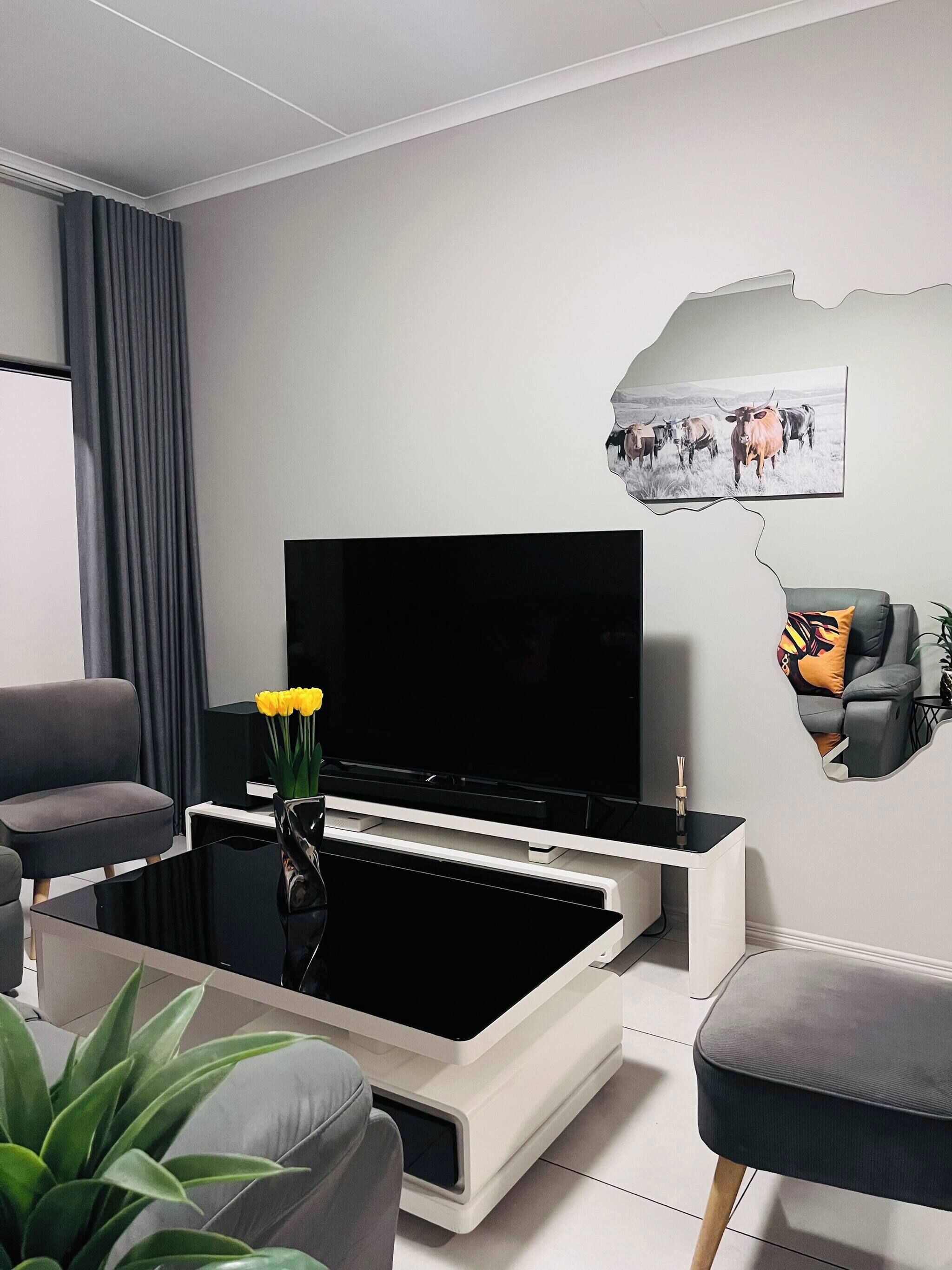 Una sala de estar moderna con un televisor de pantalla plana, una mesa de centro con un jarrón de flores amarillas, un sofá gris y un cuadro colgado en la pared de ganado.