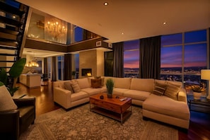 Ein modernes Wohnzimmer mit einer großen Eckcouch, einem Couchtisch und Blick auf die Stadt im Abendlicht.