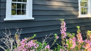 A dark-colored house with two white-framed windows and a small porch area. - Farmhouse on a River (South Kingstown)