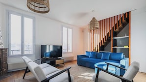 A modern living room with a sofa, two armchairs, a TV, and a staircase with wooden accents. - Magnificent Apartment -2br-6p- Saint-denis (Saint-Denis)