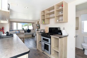 A compact kitchen with a built-in oven, a stove, and a sink. There is a seating area with a sofa and a window. - Nr Beach-30 Mins to Edinburgh-3BR Coastal Caravan (Port Seton)
