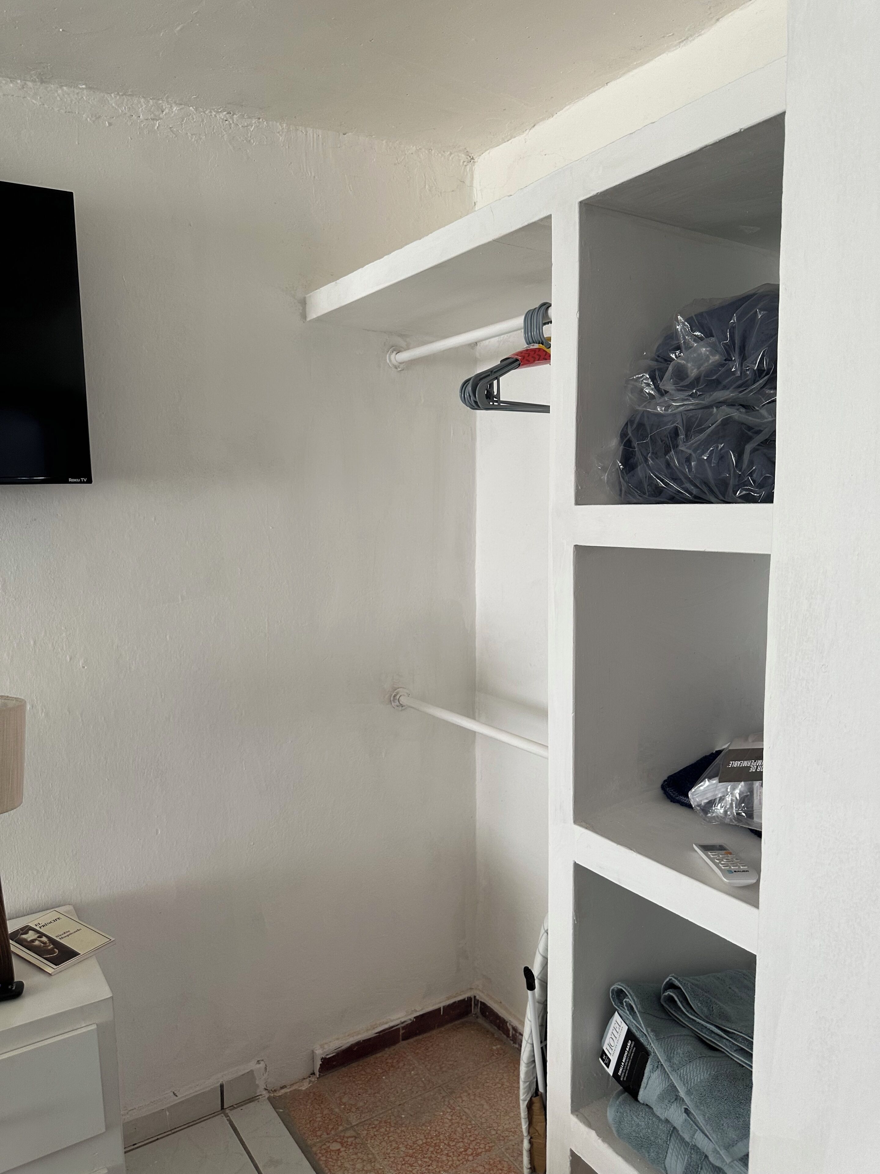 Et hjørne af et rum med hvide vægge, et fladskærms-tv monteret over et hvidt skab, og et lille bord med en lampe.