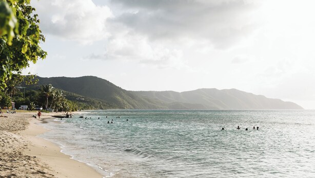 Una playa con gente nadando, una orilla arenosa y montañas al fondo.