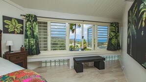 Una habitación con cama, un banco, una ventana con contraventanas, una mesita de noche y un cuadro de una planta en la pared.