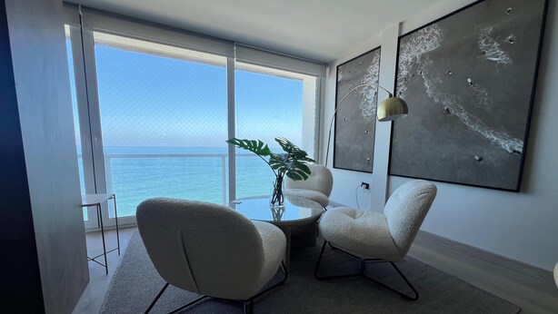 Ein modernes Wohnzimmer mit großem Fenster mit Meerblick, zwei weiße Sessel, ein Glastisch und abstrakte Wandkunst.