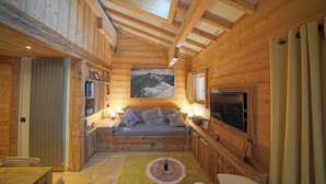 A cozy cabin interior with a wooden ceiling, a sofa with cushions, a television, and a painting of mountains.