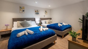 A hotel room with two beds, each with a blue blanket, a wooden nightstand, a TV, and artwork on the walls. - Cosy Battersea Flat | Zone 2 London | Sleeps 5 | Free Parking! (London)