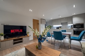Ein modernes Wohnzimmer mit Flachbildfernseher, Couchtisch aus Holz und Blumen in einer Vase.