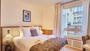 Ein Hotelzimmer mit einem Bett, einem Nachttisch, einer Lampe, einem Fenster mit Vorhängen und einem gerahmten Bild an der Wand.