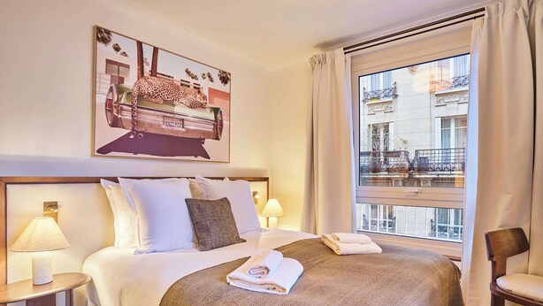 Ein Hotelzimmer mit einem großen Bett, einem Fenster mit Blick auf Gebäude, einem Gemälde über dem Bett und einem Sessel am Fenster.