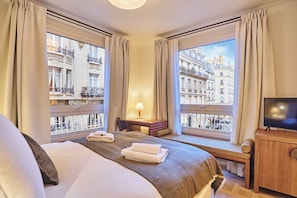 Ein Hotelzimmer mit Bett, Schreibtisch mit Lampe, Fernseher und großen Fenstern mit Vorhängen.