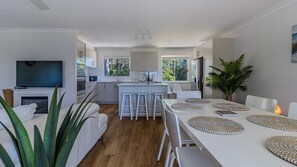 A modern kitchen with white cabinets, a central island with stools, a dining area with a white table and chairs, and a large plant. - Serene Suncrest - Pet Friendly with Heated Spa (Sussex Inlet)