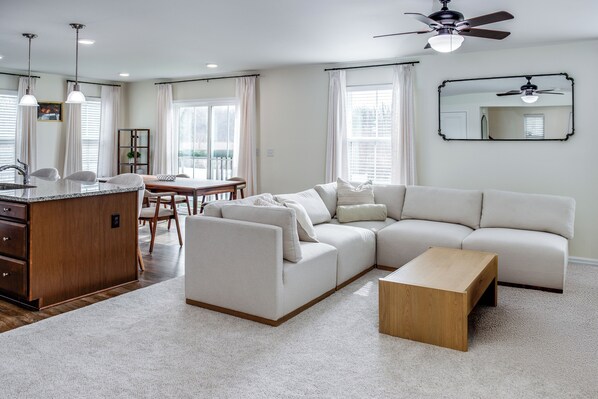 Una sala de estar moderna con un sofá modular, una mesa de centro de madera y una cocina con pileta y mesa de comedor.