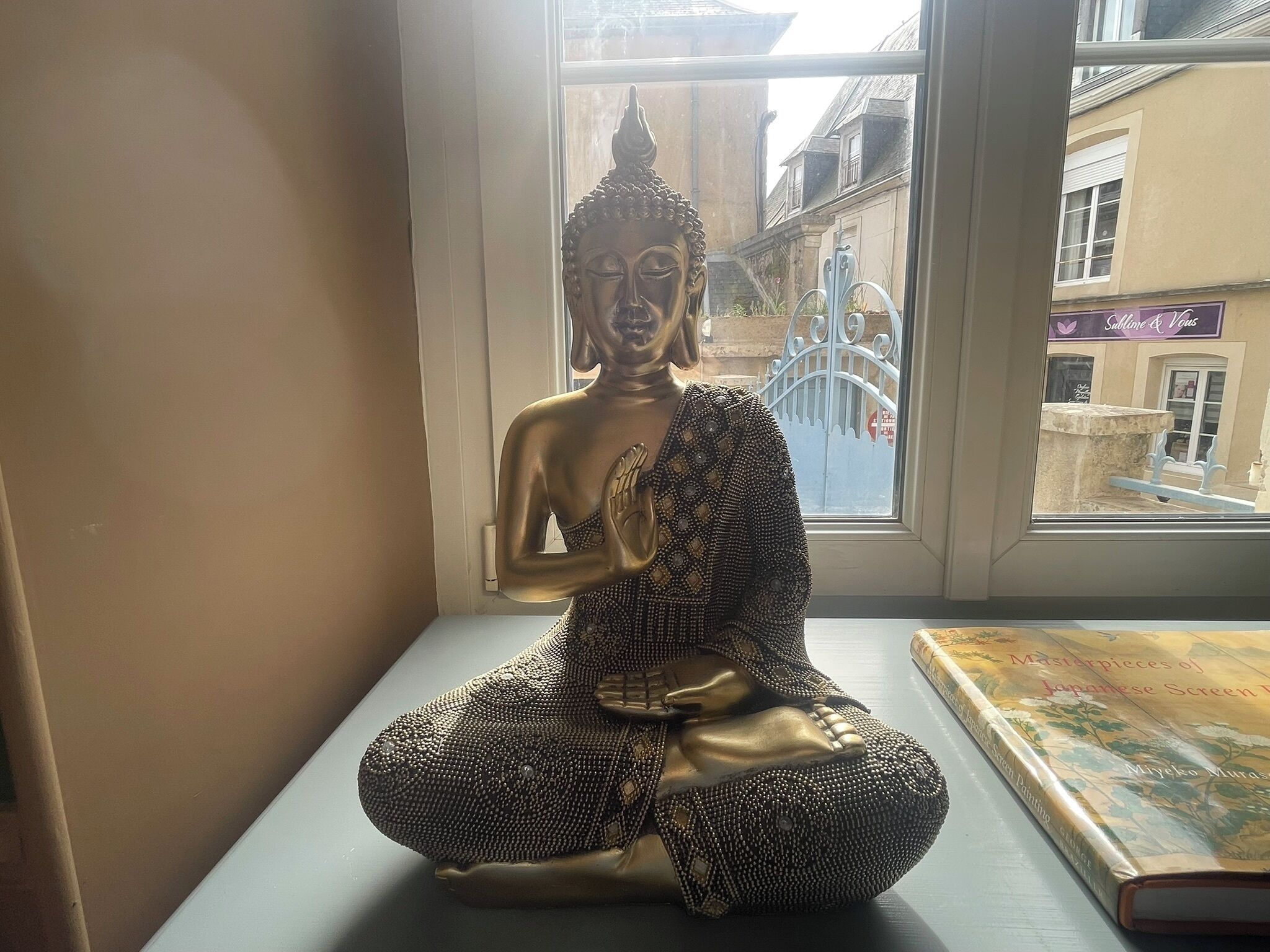 Uma estátua de Buda em um parapeito, com um livro intitulado 'Masterpieces of Japanese Screen Painting' ao lado.